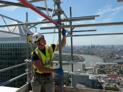 Construction worker installing scaffolding at height with city skyline in background