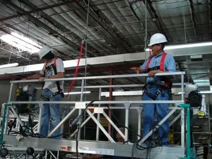 Workers using a suspended platform with safety harnesses indoors.