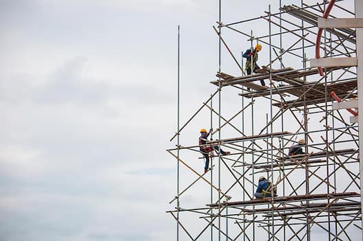 Scaffolding workers wearing safety gear on a high-rise construction site under cloudy skies.