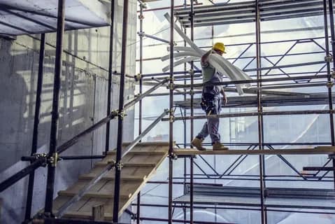 Construction worker carrying white pipes while walking on scaffolding inside a building.