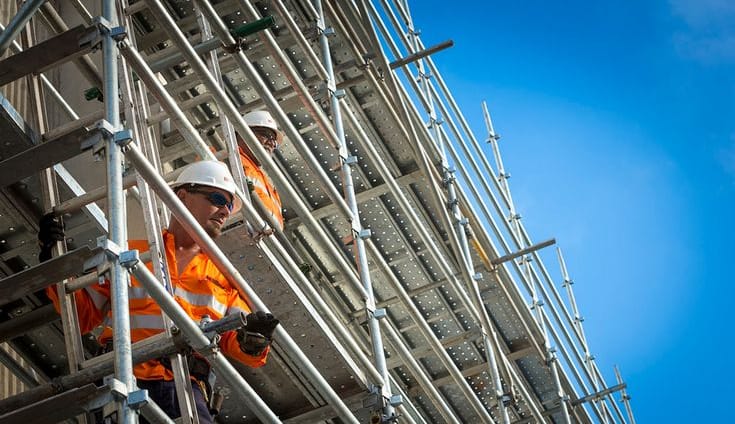 Construction workers in safety gear standing on metal scaffolding against a blue sky.
